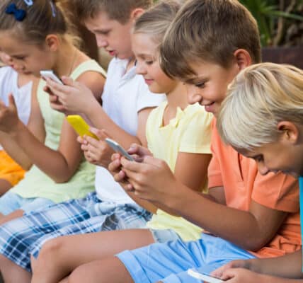 Photo of a group of young children sitting outside on a ledge all absorbed using their smartphones.