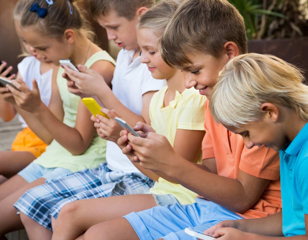 Photo of a group of young children sitting outside on a ledge all absorbed using their smartphones.