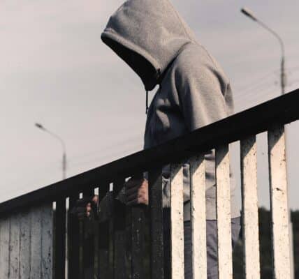 A person wearing a gray hoodie is standing on a bridge and looking down.