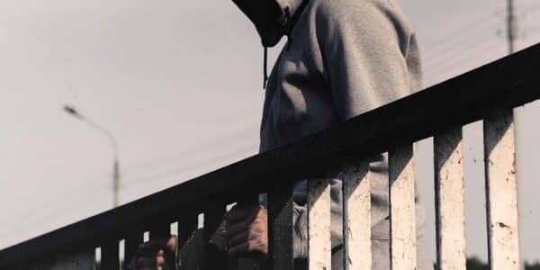 A person wearing a gray hoodie is standing on a bridge and looking down.