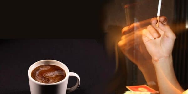 A hand holding a lit cigarette, and a cup of coffee on the table.