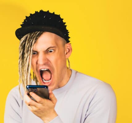 Photograph of a young man with a punk-inspired hairstyle expresses intense emotion while staring at his smartphone, highlighting the anxieties of modern communication in a vibrant, yellow backdrop.