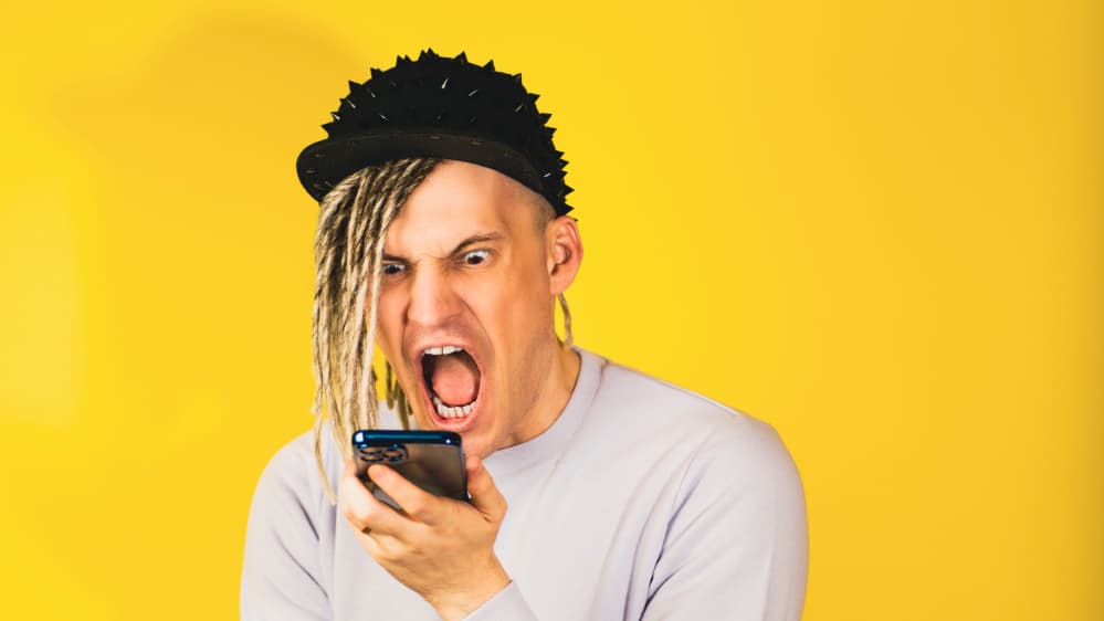 Photograph of a young man with a punk-inspired hairstyle expresses intense emotion while staring at his smartphone, highlighting the anxieties of modern communication in a vibrant, yellow backdrop.