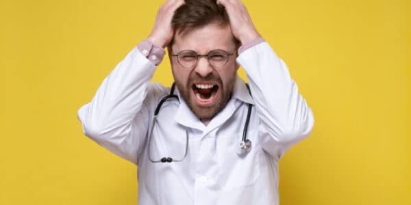 Photo of young doctor, stressed or furious, pulls his hair out and screams. Yellow background.
