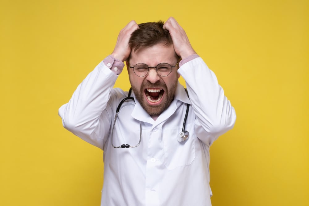Photo of young doctor, stressed or furious, pulls his hair out and screams. Yellow background.