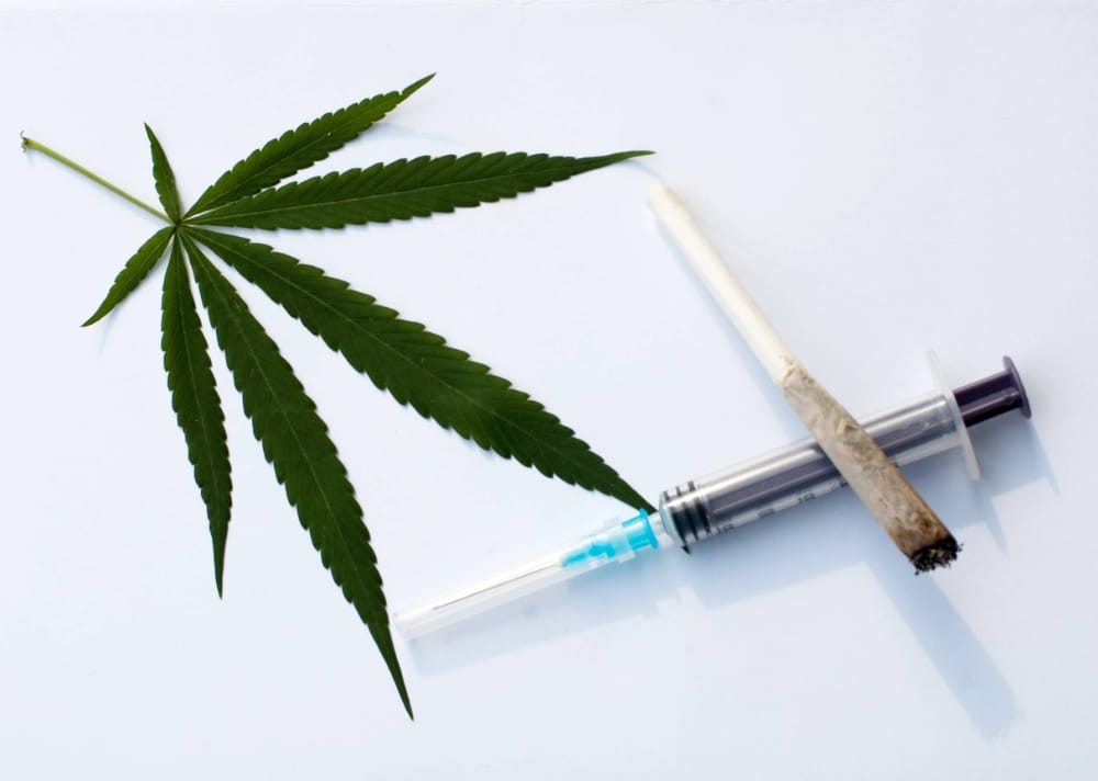 Photo of a marijuana leaf and a cannabis cigarette laying on top of an empty syringe.