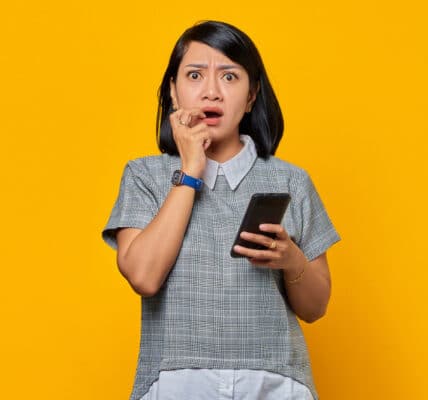 Photo of a young woman on a yellow background with a shocked expression holding a smartphone.