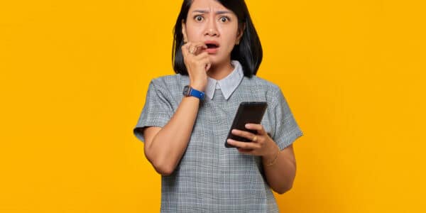 Photo of a young woman on a yellow background with a shocked expression holding a smartphone.