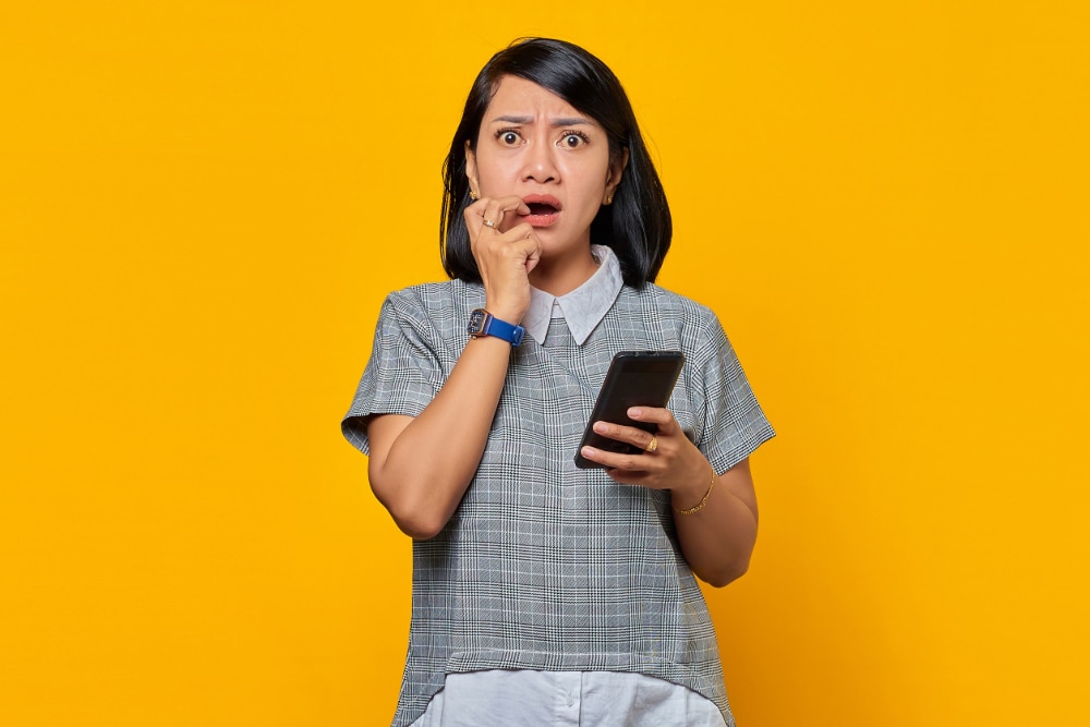 Photo of a young woman on a yellow background with a shocked expression holding a smartphone.