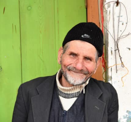 Photograph of a weathered old man with a pleasant smile against a weathered background.