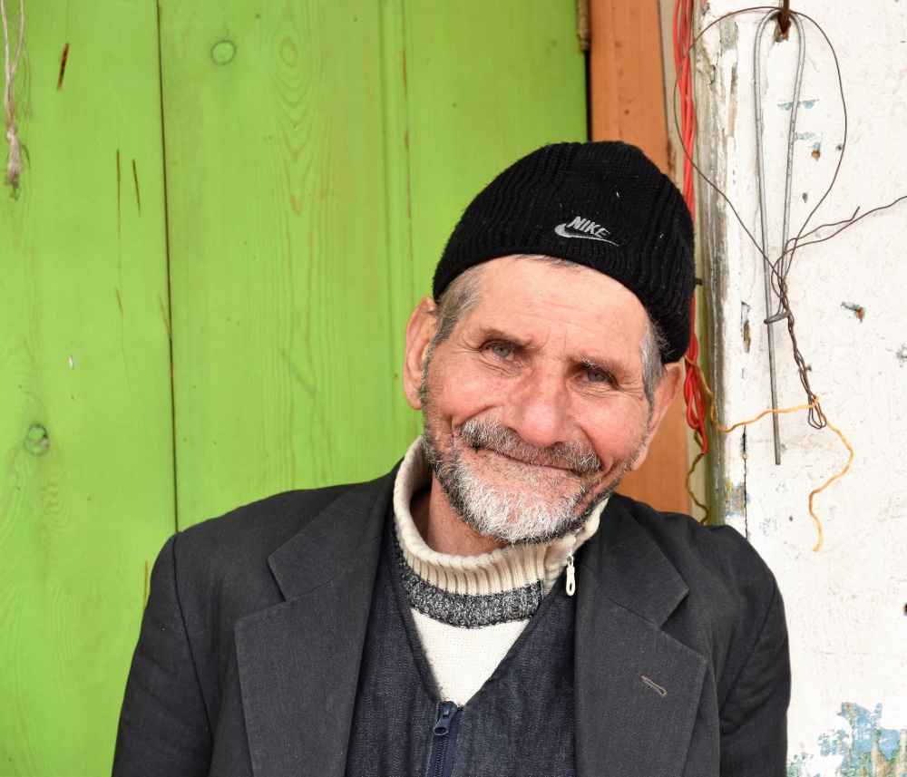 Photograph of a weathered old man with a pleasant smile against a weathered background.