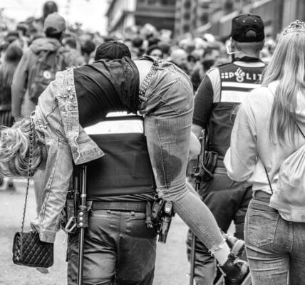 A black-and-white photo of a police officer in Amsterdam carrying a drunk young woman in a crowd.