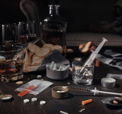 Photo of a table covered with multiple substances of abuse, including cigarettes, alcohol, powders, pills, and junk food.