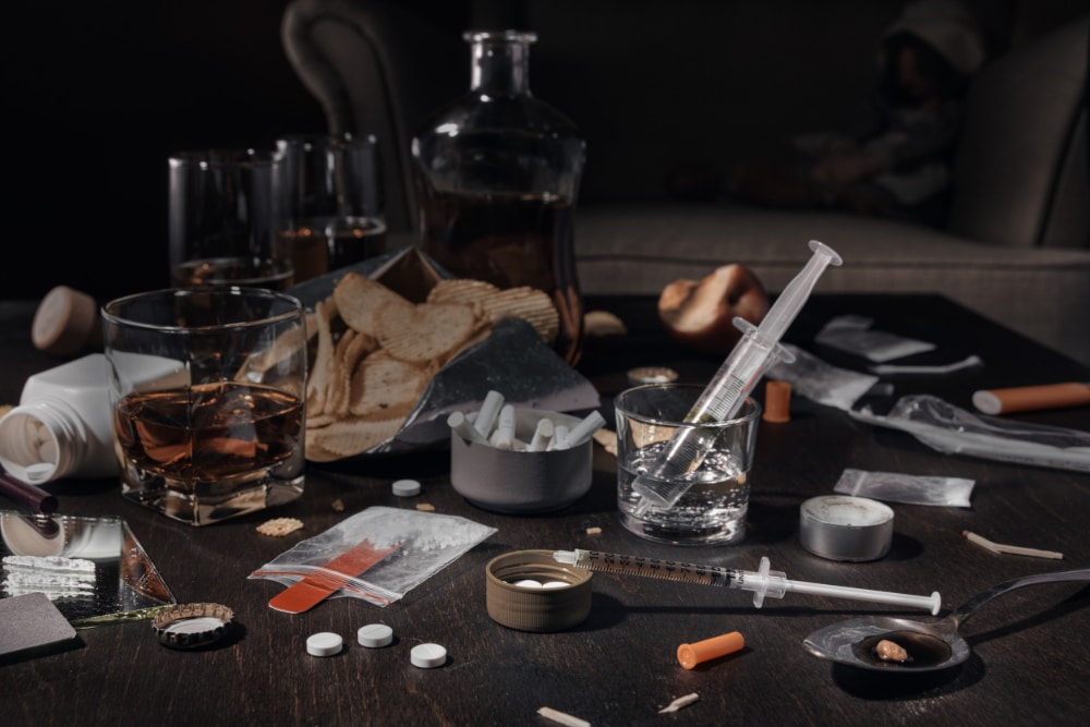 Photo of a table covered with multiple substances of abuse, including cigarettes, alcohol, powders, pills, and junk food.