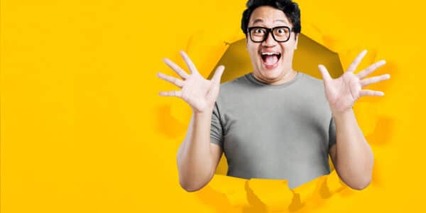 Asian man in eyeglasses with excited expression with torn paper background indicating a breakthrough.