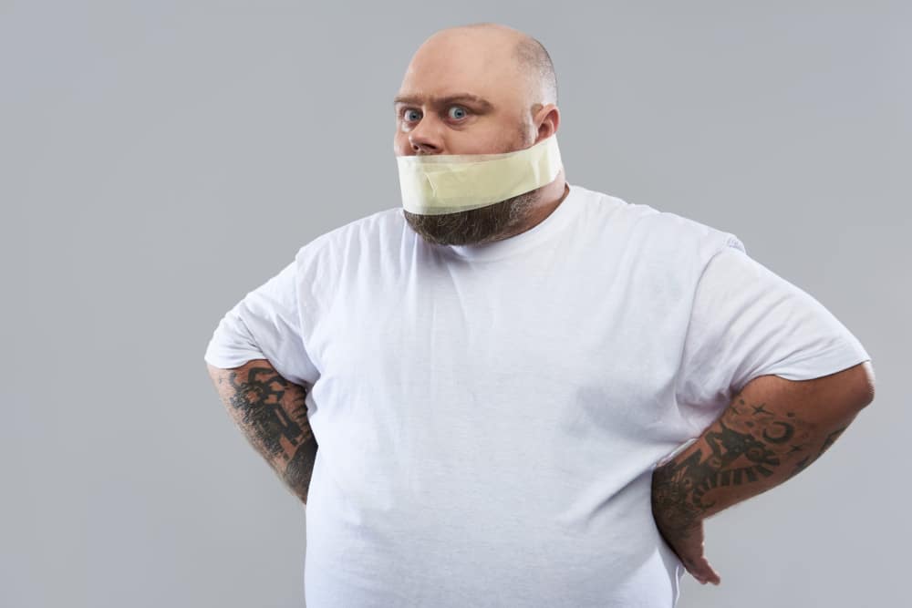 Photo of an overweight man in a white T-shirt with masking tape around his mouth as a metaphor for muting stress-related eating.