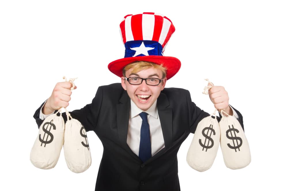 Photo of a boy in a suit and tie with an Uncle Sam top hat holding two money bags in each hand and laughing.