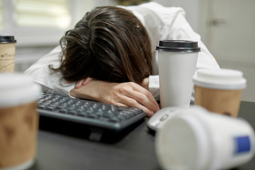 Photo of office worker asleep at her desk surrounded by coffee cups.