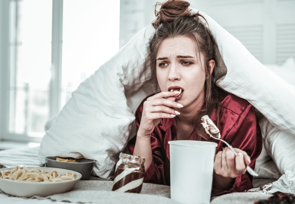 Photo of a depressed woman in bed eating ice cream and other treats.