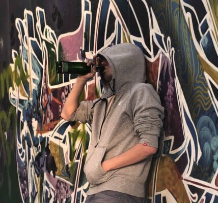 A photo of a young man wearing a hoodie drinking from the bottle against colorful background on the street.