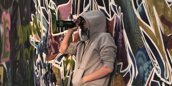 A photo of a young man wearing a hoodie drinking from the bottle against colorful background on the street.