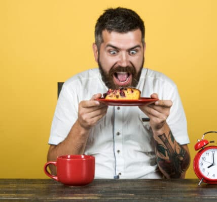 Photo of a man with an insane expression holding a plate with cake on it while an alarm clock approaches 8:00 a.m.
