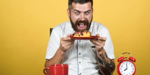 Photo of a man with an insane expression holding a plate with cake on it while an alarm clock approaches 8:00 a.m.