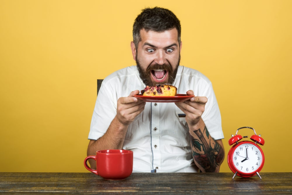 Photo of a man with an insane expression holding a plate with cake on it while an alarm clock approaches 8:00 a.m.