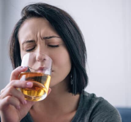 Photography of a woman with a distressed expressions drinking what looks like bourbon from a glass.