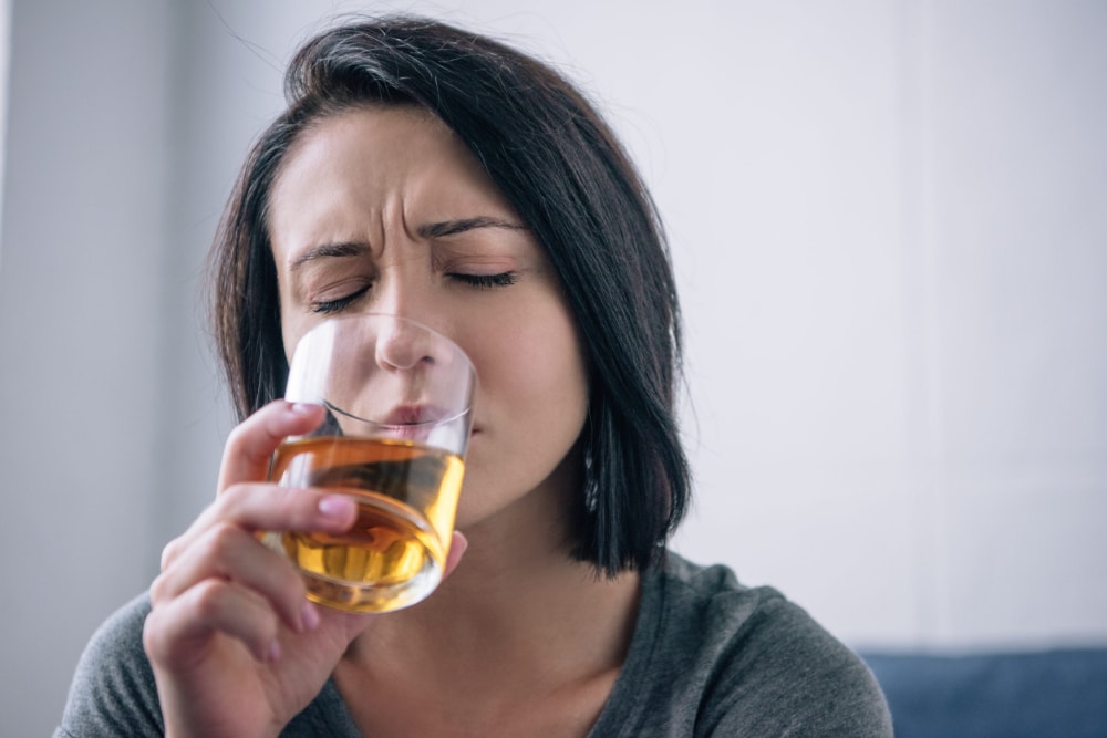 Photography of a woman with a distressed expressions drinking what looks like bourbon from a glass.