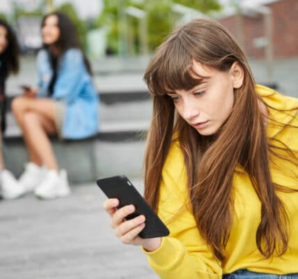 Teen girl absorbed in her smartphone as other teen girls look on.