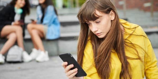 Teen girl absorbed in her smartphone as other teen girls look on.