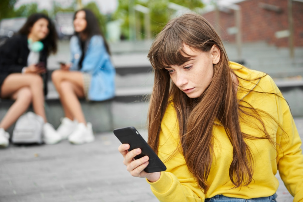 Teen girl absorbed in her smartphone as other teen girls look on.