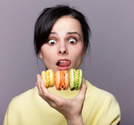 Photo of a young woman holding three macaroons in one hand with and wild look on her face.
