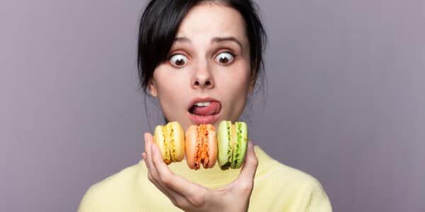 Photo of a young woman holding three macaroons in one hand with and wild look on her face.