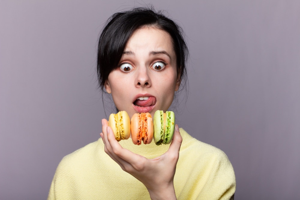 Photo of a young woman holding three macaroons in one hand with and wild look on her face.