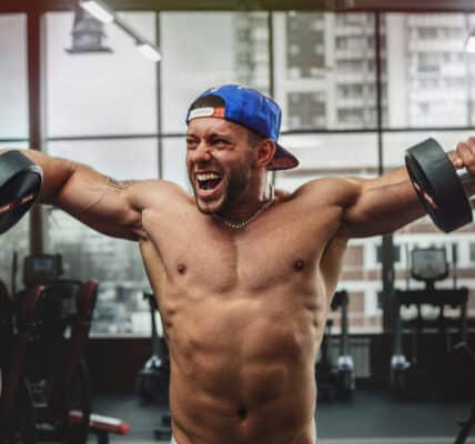 Photograph of a young body builder in a gym lifting weights with a manic look on his face.