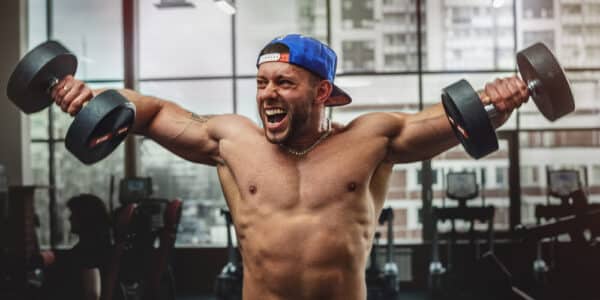Photograph of a young body builder in a gym lifting weights with a manic look on his face.