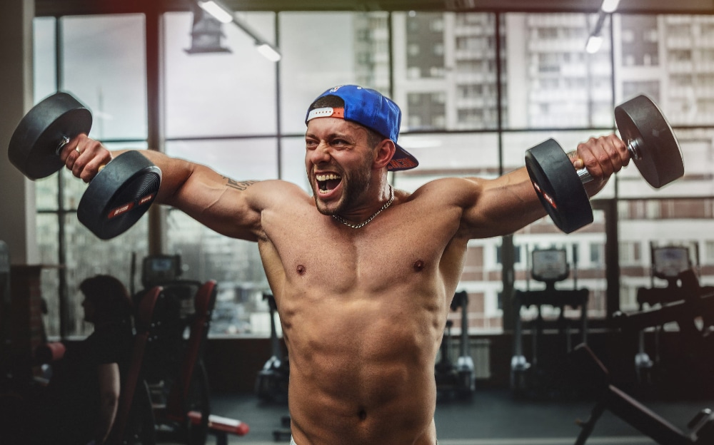 Photograph of a young body builder in a gym lifting weights with a manic look on his face.