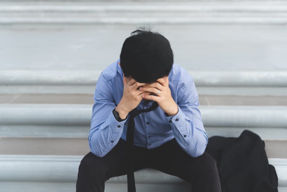 Photo of a man sitting on the steps of a building with his head in his hands in a sign of anguish.