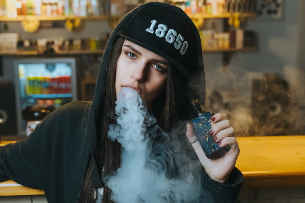 Close-up photo of a young woman in a cap smoking an electronic cigarette at the vape shop.