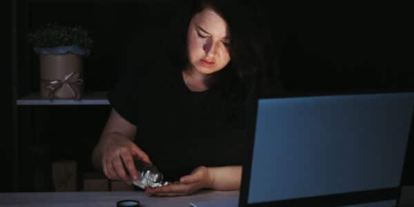 Photo of a stressed woman in front of a computer late at night taking pills out of a jar.