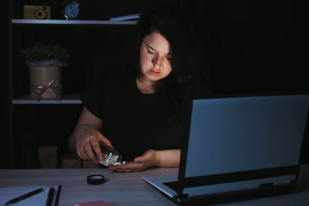 Photo of a stressed woman in front of a computer late at night taking pills out of a jar.