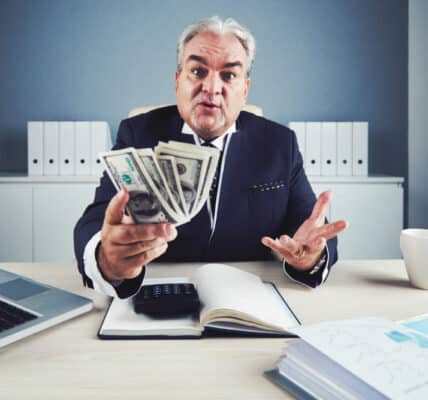Photo of a healthcare industry executive at a desk in his office with a computer open and a hand full of one hundred dollar bills.
