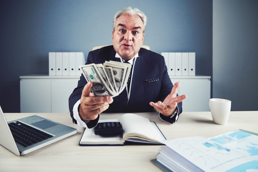 Photo of a health care industry executive at a desk in his office with a computer open and a hand full of one hundred dollar bills.