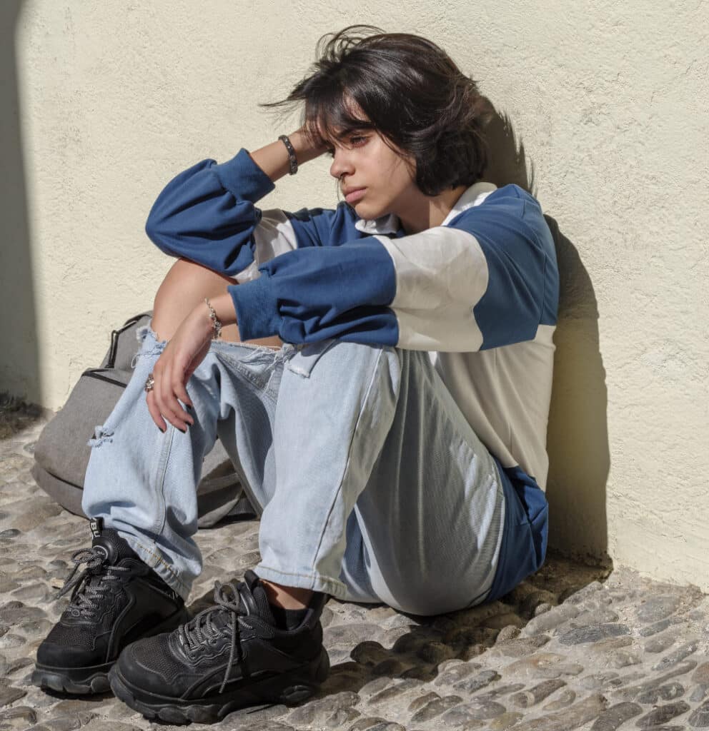 Photo of Latin teenager sitting on the ground who looks sad and distressed.