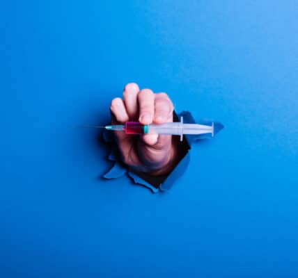 Photograph of a hand holding a GLP-1 injection bursting through a blue barrier symbolizing a breakthrough.