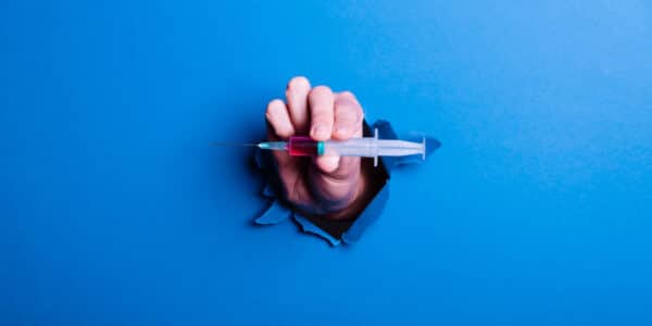 Photograph of a hand holding a GLP-1 injection bursting through a blue barrier symbolizing a breakthrough.