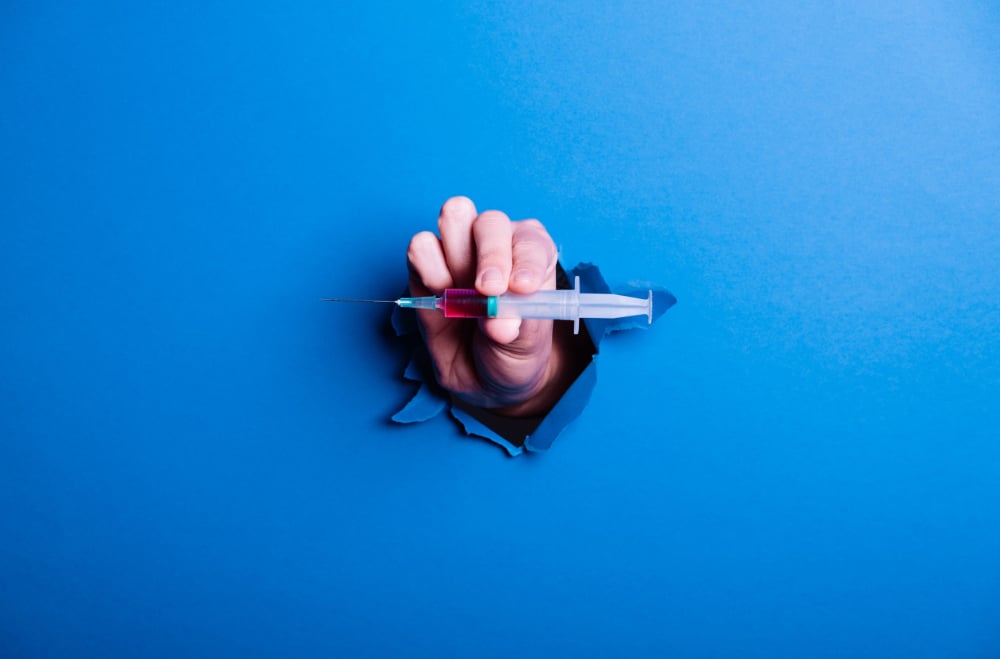 Photograph of a hand holding a GLP-1 injection bursting through a blue barrier symbolizing a breakthrough.